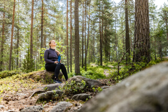 Nainen istuu kivellä juomapullon kanssa keskellä vihreää mäntykangasta