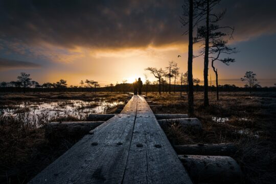 Kaksi ihmistä Linnaistensuon pitkospuilla auringon paistaessa horisontissa