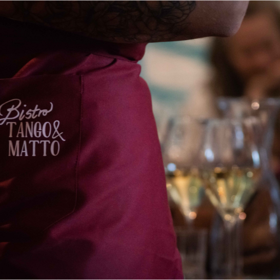 Bistro Tango & Matto Heinola, tarjoilijan essu etualalla, ruokavieraiden ja viinilasien hahmoja taustalla